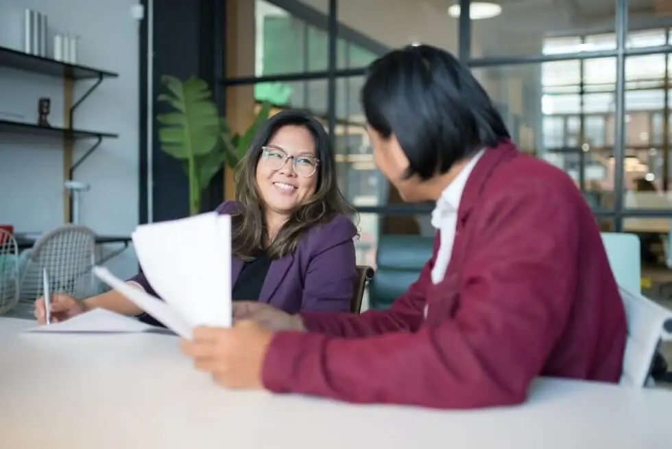 Smiling Asian woman sitting at a desk with colleague holding quarterly performance reviews