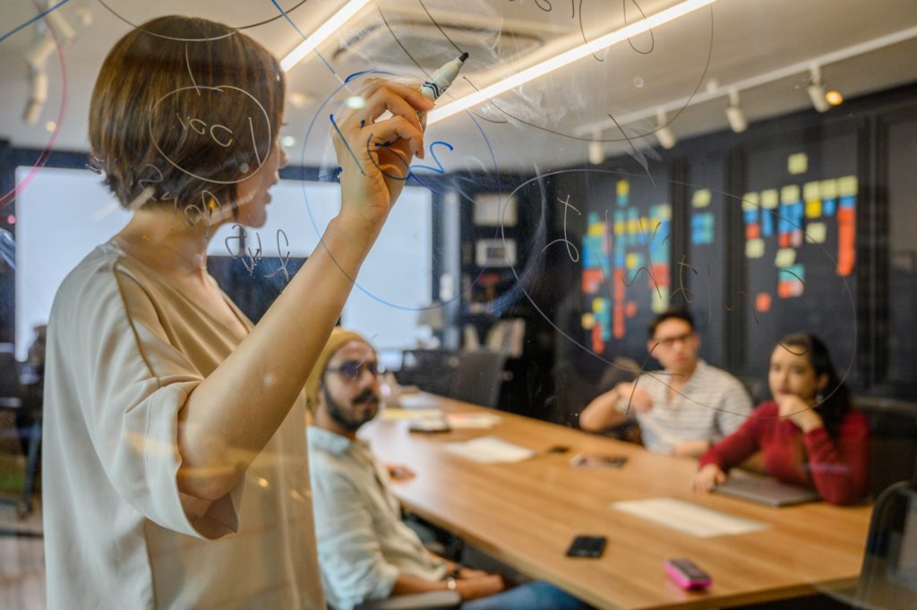Woman writing on glass about effective management with colleagues sitting